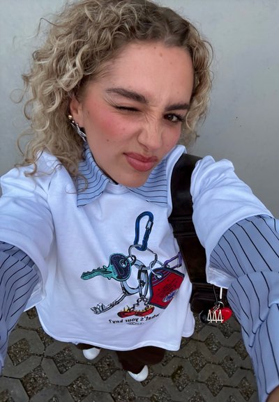 Camiseta blanca con un gráfico de llave colorido, cuello a rayas azules y pantalones de pierna ancha marrones. Llaverito con un corazón rojo y un emblema plateado visible.