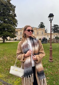 Γυναίκα με γυαλιά ηλίου, καμηλό παλτό και πολύχρωμο κασκόλ στέκεται σε πράσινο γκαζόν κοντά σε ιστορικό κτίριο και παλιά λαμπτήρα δρόμου.