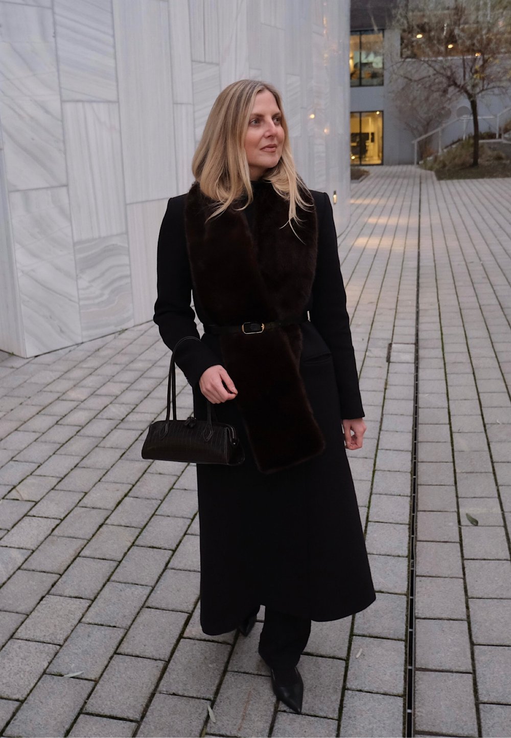 Un long manteau noir avec une écharpe en fourrure marron, ceinturé à la taille, accompagné d'un petit sac à main noir et de chaussures noires pointues.
