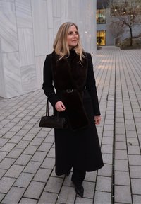 Un long manteau noir avec une écharpe en fourrure marron, ceinturé à la taille, accompagné d'un petit sac à main noir et de chaussures noires pointues.