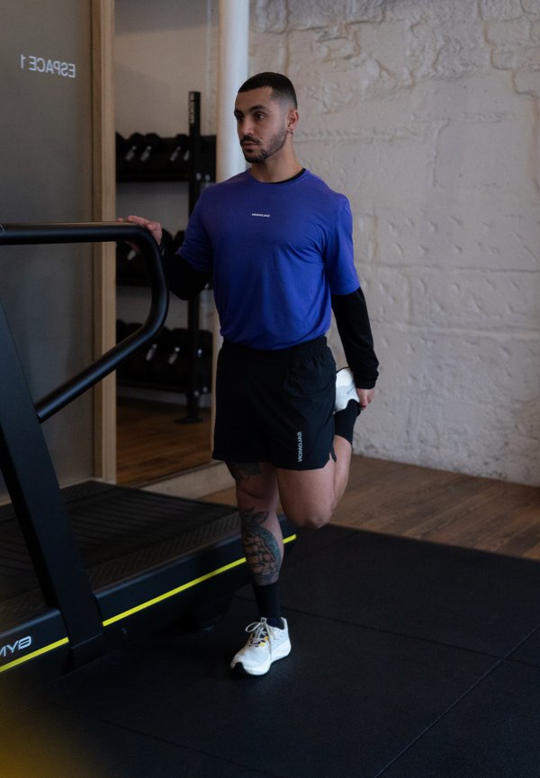 Mann i blå skjorte og svarte shorts strekker quadricepsmuskelen ved å holde foten bak seg, stående ved siden av et tredemølle i treningsstudio.