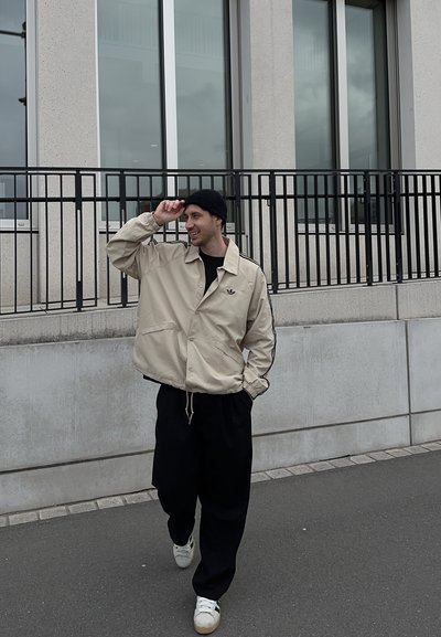 Hombre vistiendo chaqueta beige, pantalones negros y gorro, caminando al aire libre junto a un edificio de concreto con grandes ventanas y una barandilla de metal negra.