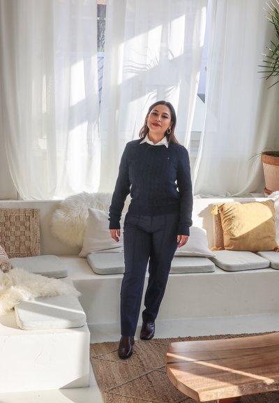 Suéter de punto trenzado navy sobre una camisa blanca con cuello, combinado con pantalones oscuros y botas marrones hasta el tobillo, de pie en una habitación iluminada.