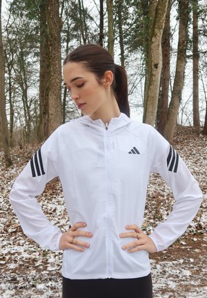 Chaqueta deportiva ligera blanca con capucha, cremallera frontal y acentos negros de tres rayas en las mangas. Textura suave.