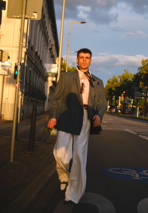 Ein Mann, der einen übergroßen Blazer und weite weiße Hosen trägt, geht bei Sonnenuntergang mit einer weißen Rose auf der Stadtstraße.