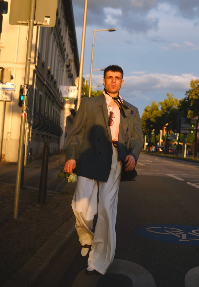 Man wearing oversized blazer and wide white pants walks on city street holding a white rose at sunset.