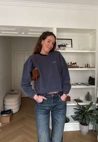 Femme aux longs cheveux bruns portant un sweat-shirt bleu marine et un jean, les mains dans les poches, debout à l'intérieur près d'étagères avec des décorations et des plantes.