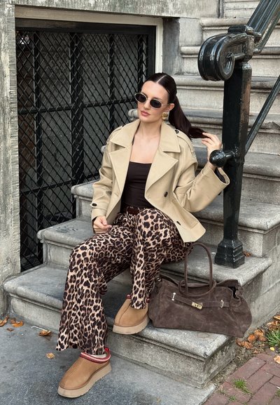 Abrigo trench beige, camiseta sin mangas marrón, pantalones de leopardo marrón, zapatos de felpa color beige, bolso de ante marrón, gafas de sol ovaladas, cabello largo y oscuro.