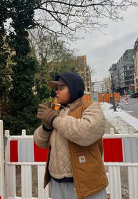 Καπιτονέ μπεζ και καφέ μπουφάν με μόνωση, μαύρη κουκούλα, πλεκτά γάντια, στέκεται κοντά σε κατασκευαστικά εμπόδια, με αστικό φόντο και δέντρα.