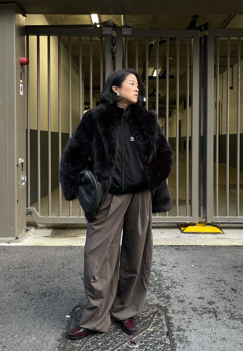 Une femme se tient sur le trottoir devant une grille en métal, portant une veste en fourrure noire, un sweat à zip Adidas sombre, un pantalon large gris, et tenant un sac noir tissé.