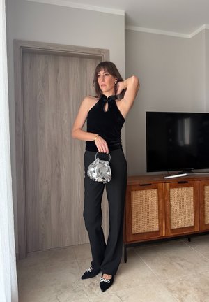 Mujer con top sin mangas negro y pantalones, sosteniendo un bolso de lentejuelas plateado, de pie junto a un armario de madera con un televisor.