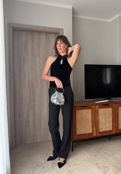 Mujer con top sin mangas negro y pantalones, sosteniendo un bolso de lentejuelas plateado, de pie junto a un armario de madera con un televisor.