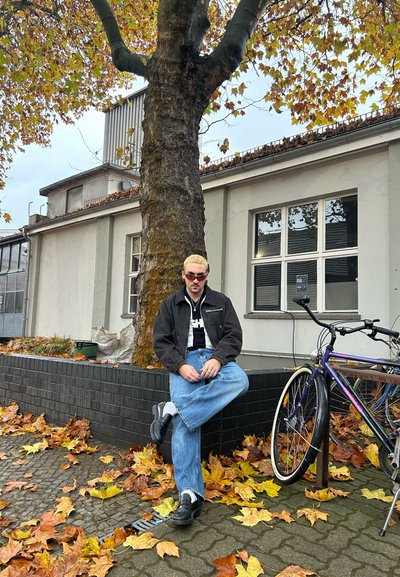 Persona con chaqueta negra, camisa blanca y jeans azules sentado junto a un árbol. Hojas amarillas caídas rodean una bicicleta apoyada cerca.