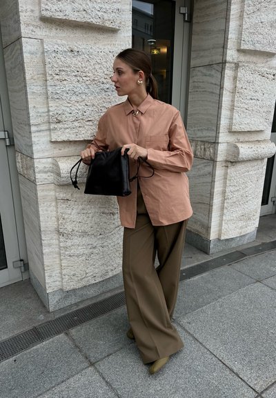 Camisa de botones de color rosa claro con cuello, de corte oversize, combinada con pantalones anchos marrones y una mochila negra con cordón.