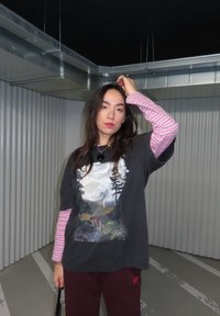 Femme aux longs cheveux noirs portant un t-shirt graphique noir superposé à un t-shirt à manches longues rose rayé, debout dans un espace industriel intérieur avec des conduits métalliques.