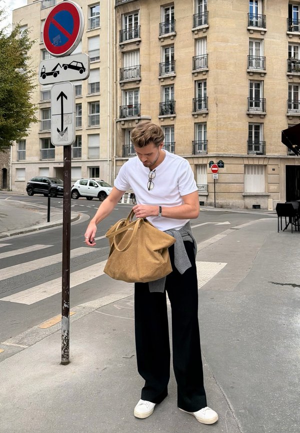 Hombre con camisa blanca y pantalones negros de pie en la acera junto a señales de prohibido aparcar y zona de remolque, sosteniendo una gran bolsa marrón claro.