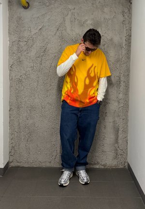 Camiseta amarilla con un patrón de llamas en la parte inferior, combinada con jeans azules y zapatillas plateadas. El fondo presenta paredes grises texturizadas.