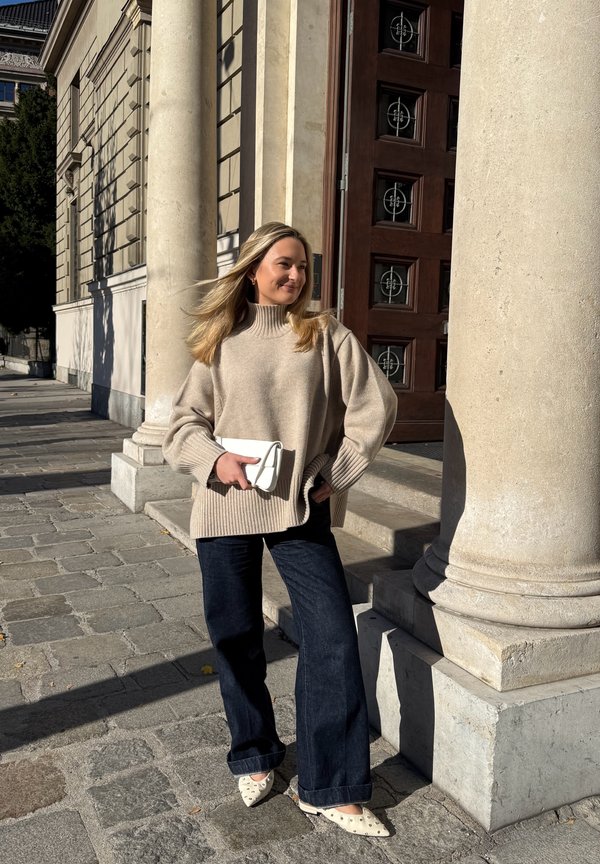 Beige oversized sweater med ribbet manchetter, mørke denim-flare jeans og hvid clutch. Spidse hvide sko med små nitter. Stenkolonner i baggrunden.