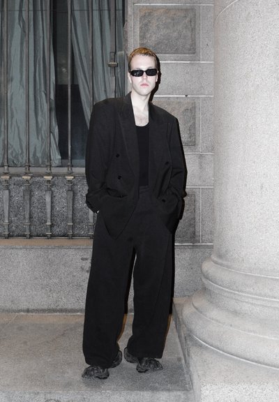 Traje negro oversize con chaqueta de doble botonadura, top negro sin mangas y pantalones anchos, combinado con gafas de sol negras y zapatos negros de suela gruesa.