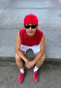 Κόκκινο σκουφί, αμάνικο κόκκινο μπλουζάκι, λευκές βερμούδες και κόκκινα αθλητικά παπούτσια με λευκές ρίγες. Άτομο καθισμένο σε μια πέτρινη επιφάνεια, με τα χέρια ενωμένα.