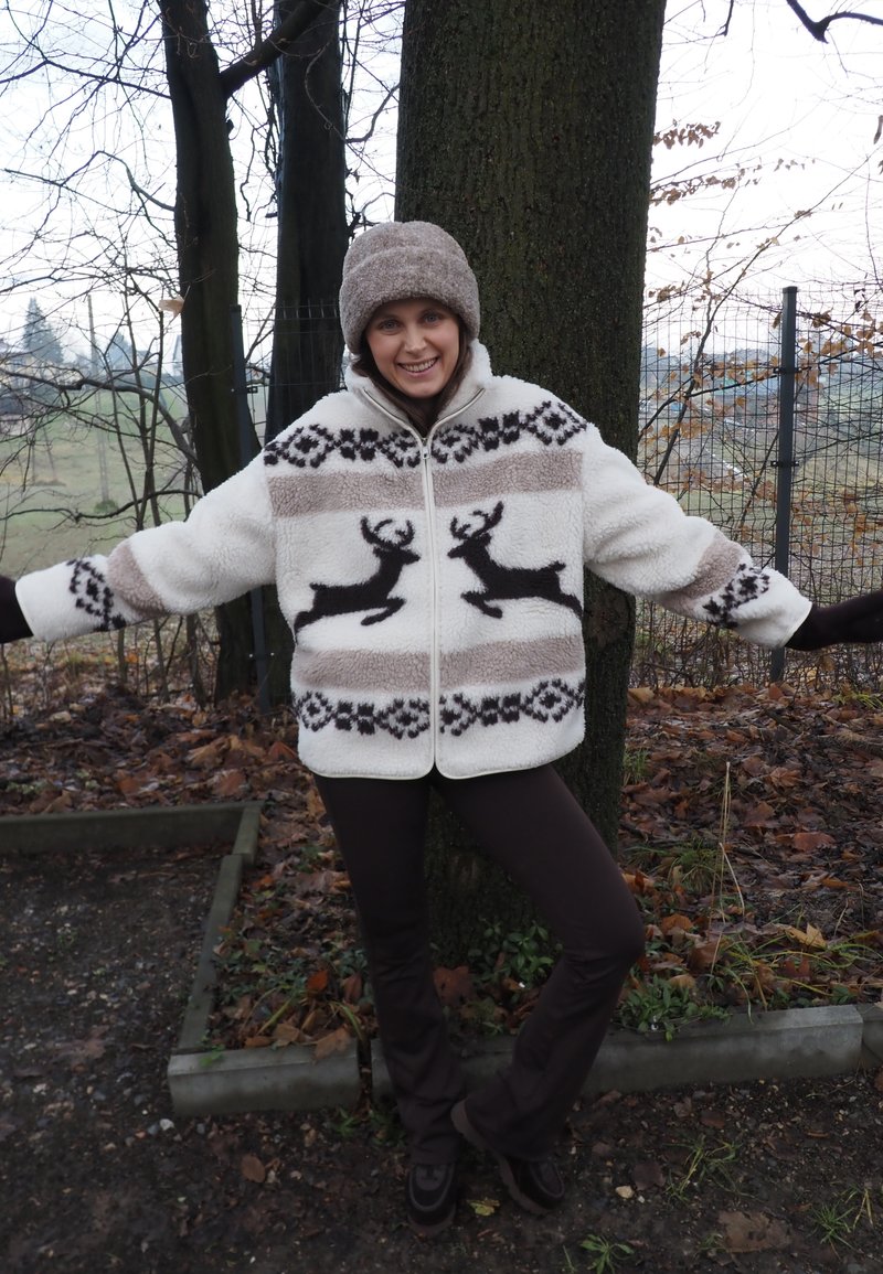 Veste blanche duveteuse avec motifs de rennes noirs et accents bruns, assortie à un chapeau brun et un pantalon brun, posant près d’un arbre.