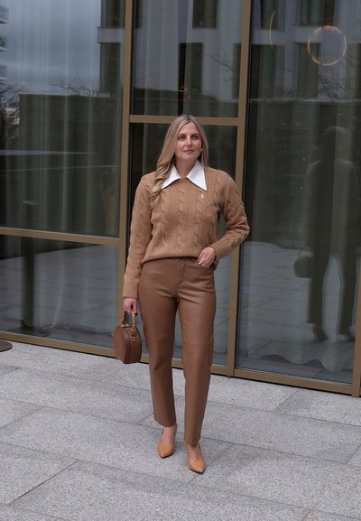 Suéter de punto color canela con cuello blanco, combinado con pantalones de cuero marrón ajustados y tacones de punta en tono marrón claro, sosteniendo un bolso redondo.