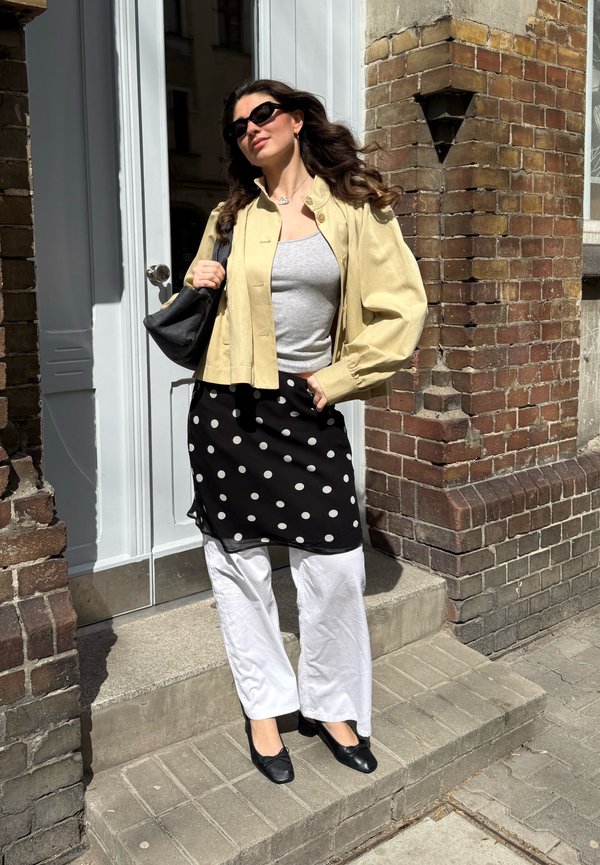 Femme portant des lunettes de soleil, veste beige, haut gris, jupe noire à pois portée sur un pantalon blanc et chaussures noires, debout sur des marches extérieures.