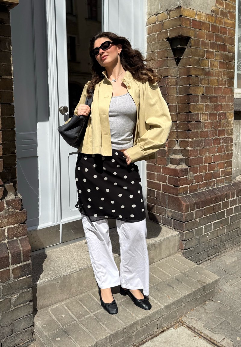 Γυναίκα με γυαλιά ηλίου, μπεζ μπουφάν, γκρι τοπ, μαύρη φούστα με πουά πάνω από λευκό παντελόνι και μαύρα παπούτσια, που στέκεται σε υπαίθρια σκαλιά.