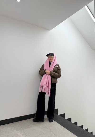 Chaqueta acolchada marrón, pantalones deportivos negros y una bufanda grande rosa con flecos, de pie en un espacio minimalista bien iluminado con suelo gris.