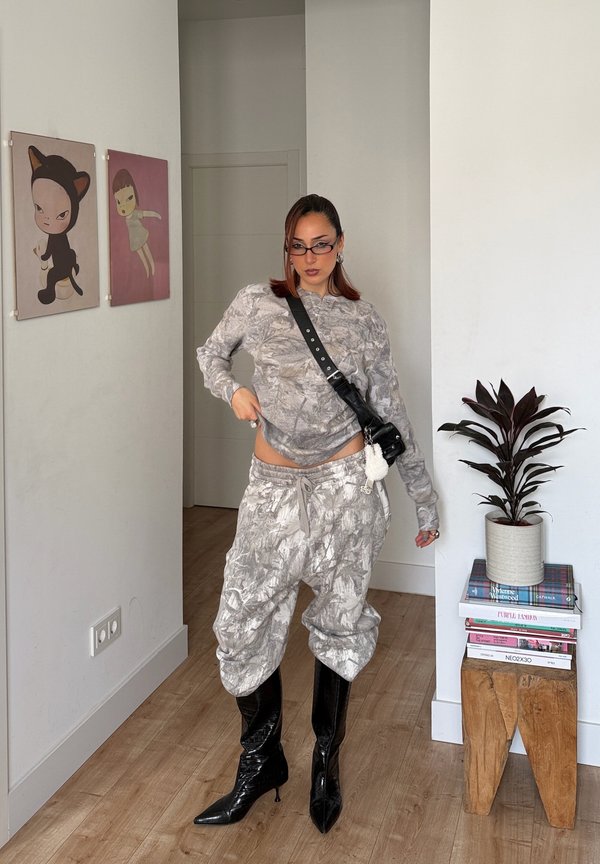 Oseba v sivi obleki z vzorcem in hlačah, črnih čevljih z visokimi petami ter majhno torbico čez ramo, stoji v sobi z umetniškimi deli in rastlino na knjigah.