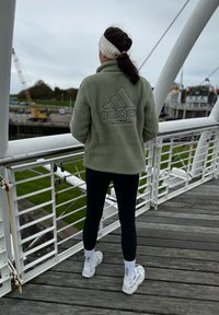 Veste en polaire verte avec le logo "Adidas", leggings noirs et baskets blanches. Cheveux attachés avec un bandeau blanc, debout sur une passerelle en bois.