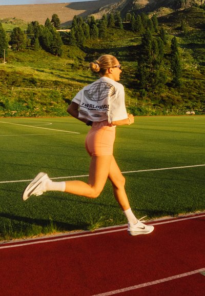 Persona corriendo en una pista, vistiendo una camiseta corta blanca con un diseño gráfico, pantalones cortos melocotón y zapatillas deportivas blancas con detalles en negro.