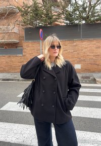 Femme aux cheveux blonds, portant des lunettes de soleil, un manteau noir croisé et un sac à franges, debout sur un passage piéton près d'un mur en briques.