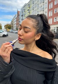 Pomadka w srebrnej tubce nałożona na usta. Obiekt nosi czarny, żeberkowany top bez ramiączek i ma długie, falowane włosy. Tło miejskie.