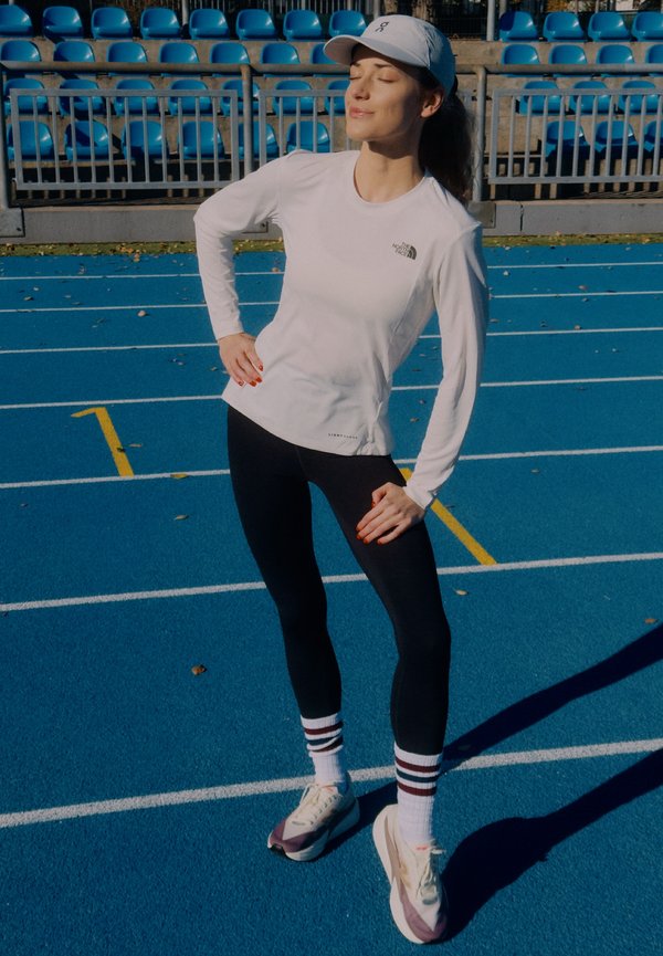 Sportininkas apsirengęs baltu marškinėlių ilgomis rankovėmis, juodomis tamprėmis ir baltais sportbačiais su violetiniais akcentais, stovi ant mėlyno bėgimo takelio.