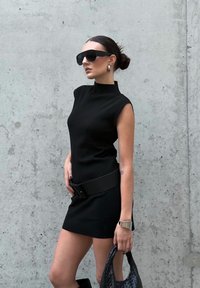 Zwarte mouwloze jurk met een hoge halslijn en een nauwsluitend ontwerp, taille gepint met een bijpassende riem. Model is voorzien van oversized zonnebril.