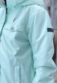 Ανοιχτό μέντα πράσινο μπουφάν από απαλό, ελαφρώς γυαλιστερό ύφασμα, με φερμουάρ, κουκούλα, μπροστινή τσέπη και υφή μαύρη πατσίτσα στον ώμο.