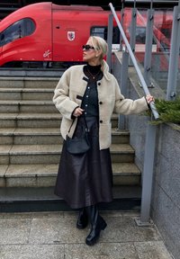 Giacca oversized in finta pelliccia shearling color crema con bottoni neri, indossata sopra un maglione scuro e una lunga gonna di pelle marrone; borsa a spalla nera; stivali neri.