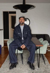 Donkerblauwe denimjack en -broek met witte stiksels, grijs t-shirt, zwarte schoenen, zittend op groene stoelen aan een houten tafel onder een zwarte lamp.