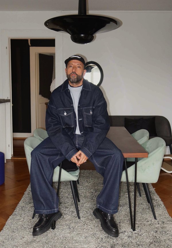 Donkerblauwe spijkerjas en broek met witte stiksels, grijs t-shirt, zwarte schoenen, zittend op groene stoelen aan een houten tafel onder een zwarte lamp.
