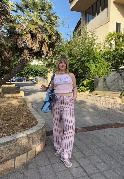 Persona que lleva una blusa corta de color rosa claro con texto de logo azul, pantalones de tiro ancho a rayas en rosa y blanco, y sandalias, sosteniendo una bolsa de tote azul.