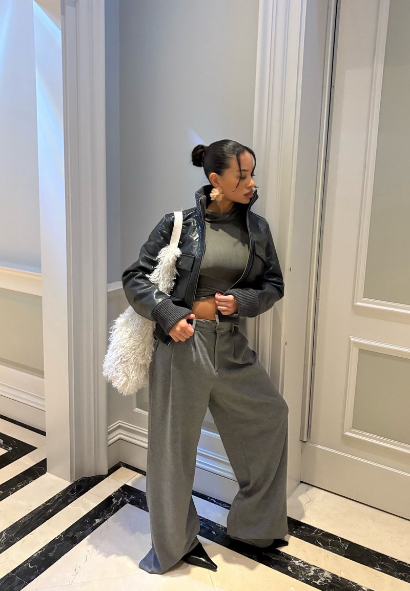 Veste noire en simili cuir, haut court gris, pantalons larges gris, sac à main blanc duveteux, chaussures noires élégantes, fond clair avec sol géométrique.