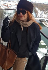 Femme portant un manteau noir, un chapeau marron, des lunettes de soleil et des gants, se tenant sur un balcon surplombant une rivière enneigée et le paysage urbain.