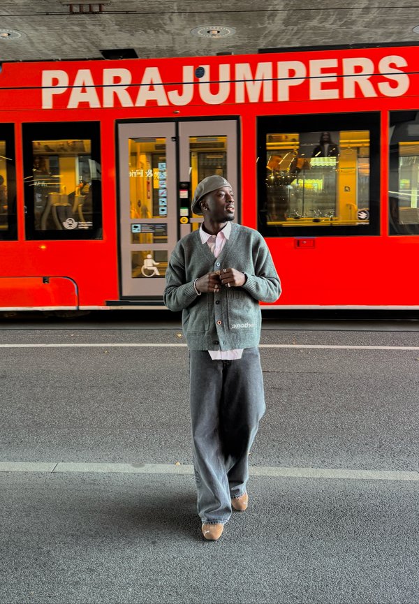 Un cardigan tricoté vert sur une chemise rose clair, un pantalon large gris et des chaussures beiges ; un tram rouge avec le texte "PARAJUMPERS" en arrière-plan.