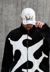 Juoda ir balta pūkuota užtrauktuko striukė su abstrakčiu dizainu; dėvima balta kepurė su spausdintu erelio motyvu, šiek tiek pakreipta.