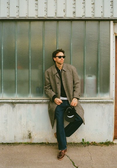 Abrigo de espiga gris sobre un suéter negro, jeans azules y zapatos marrones. Sosteniendo una gorra negra, gafas de sol, con un fondo urbano texturizado.