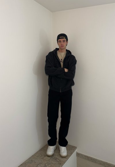 Sudadera con cremallera negra, camiseta crema con estampado de "A", pantalones negros y zapatillas blancas; de pie contra una pared lisa.