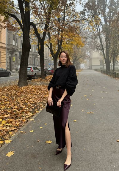 Suéter negro texturizado, falda de satén burdeos con abertura lateral, tacones puntiagudos burdeos y un bolso clutch negro. Hojas de otoño en el suelo.