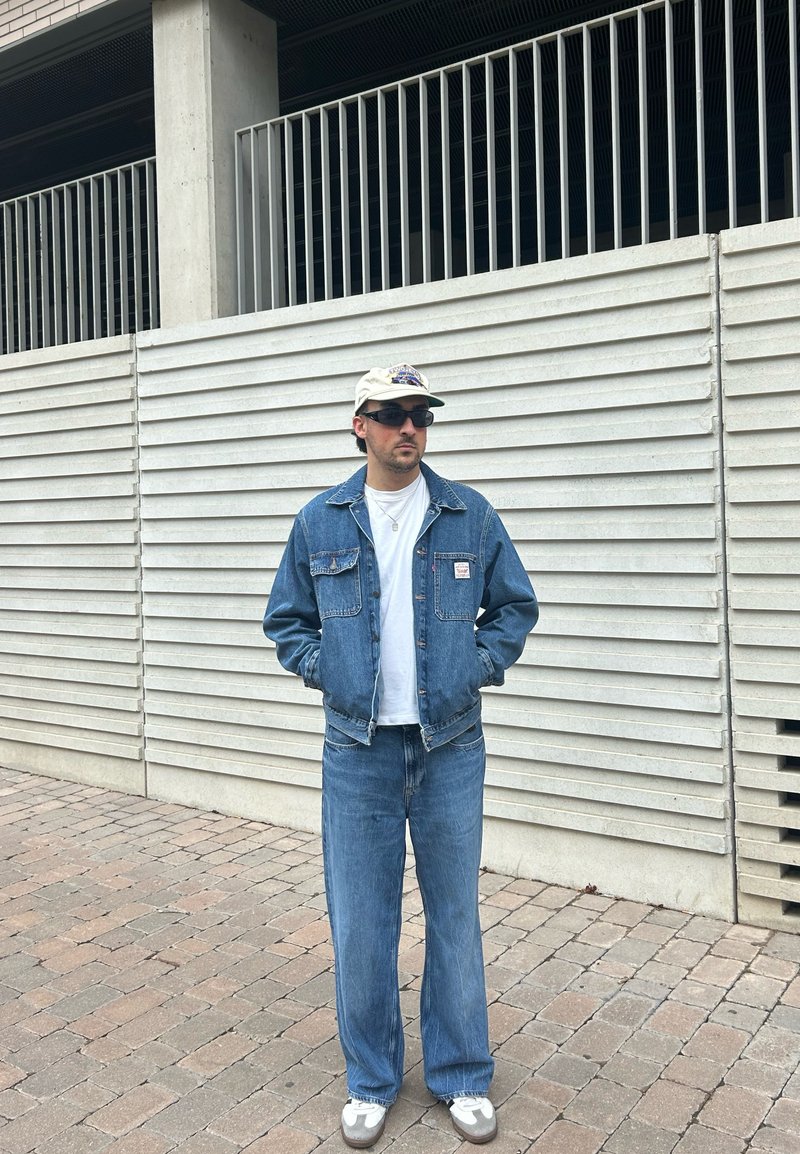 Jeans jacket και ταιριαστά παντελόνια σε ανοιχτό μπλε, συνδυασμένα με λευκό t-shirt και μπεζ καπέλο. Οι γυαλιά ηλίου ολοκληρώνουν το casual look.
