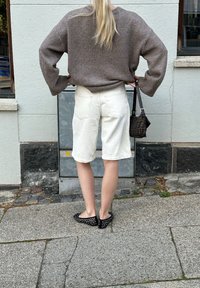 Γκρι φούτερ υπερμεγέθους, λευκά σόρτς, μαύρες φλατ παπούτσια με πουά. Η τσάντα έχει καφέ σχέδιο. Το φόντο περιλαμβάνει έναν υφασμένο τοίχο και πλακόστρωτο.